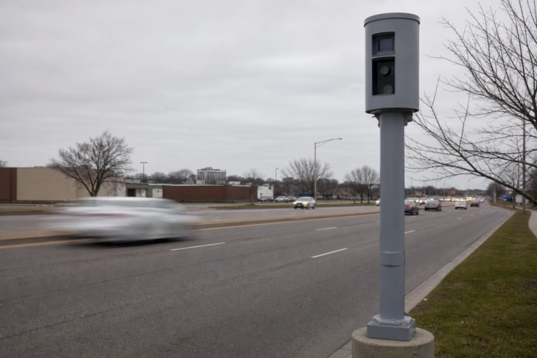 Automated speed camera pole beside a wide urban arterial street with passing cars