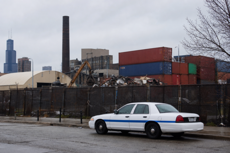 Chicago industrial block with a scrapyard fence and smokestack, a city sedan parked at the curb