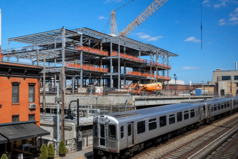 An elevated train station under construction with a train passing under a blue sky