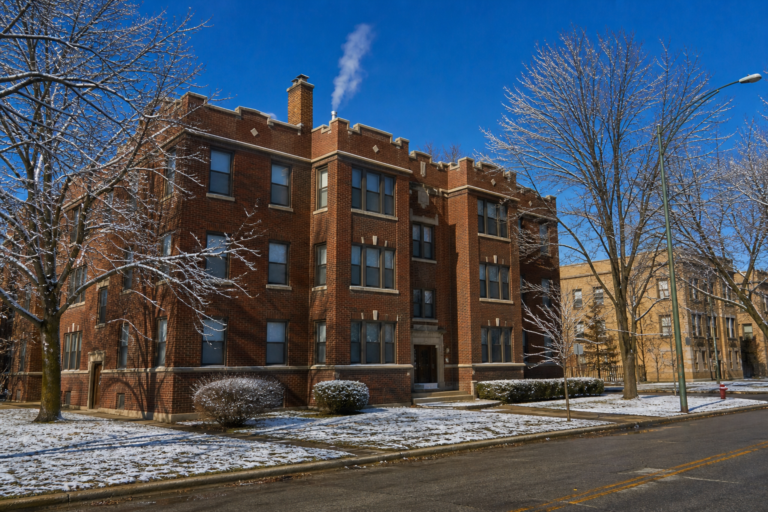 A three-story brick courtyard apartment building on a clear winter morning, light snow on the lawn