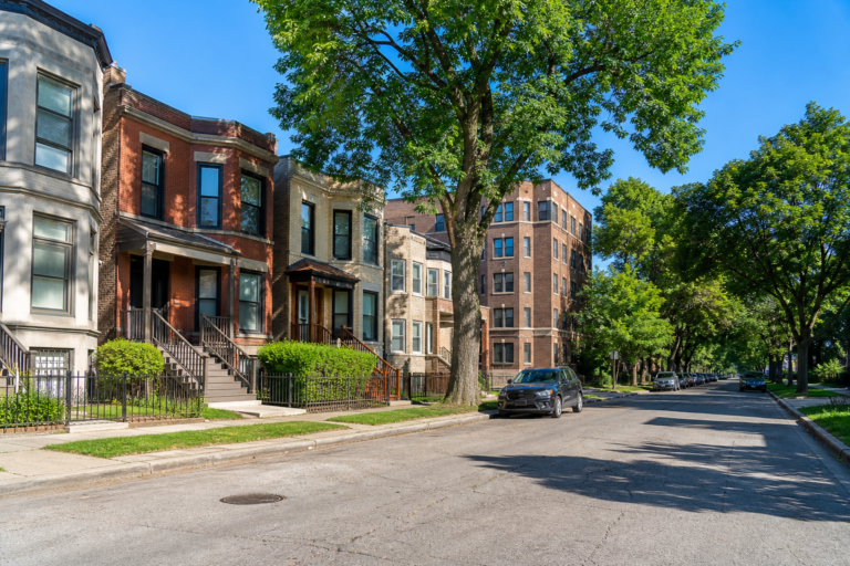 Greystone and brick rowhouses with bay windows under tall shade trees on a level street