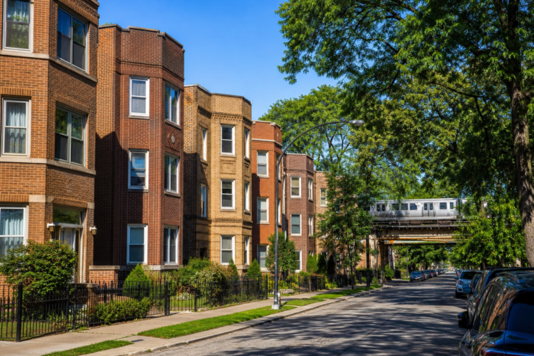 Brick two-flats and three-flats with bay windows on a level street, elevated train tracks beyond